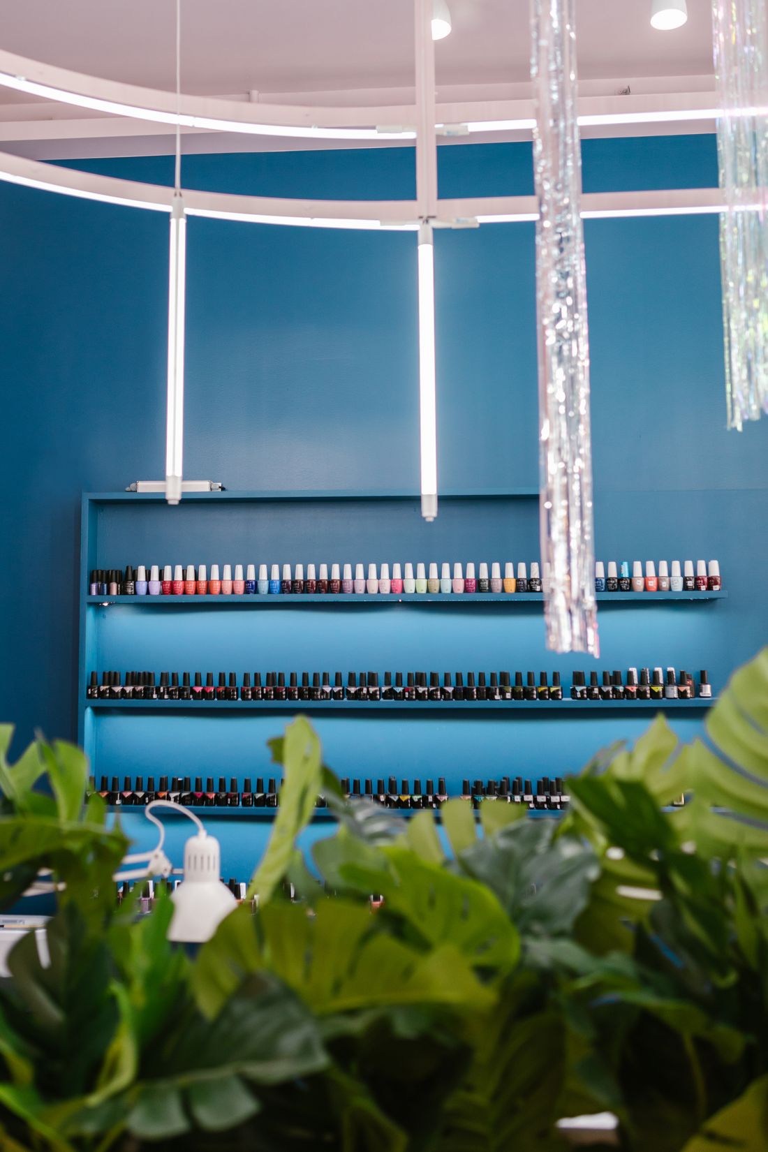 A deep-blue wall with a long shelf of gel-polish bottles in rows, white ring-light tubes and iridescent tinsel hanging in front, and big green leaves blurred in the foreground.