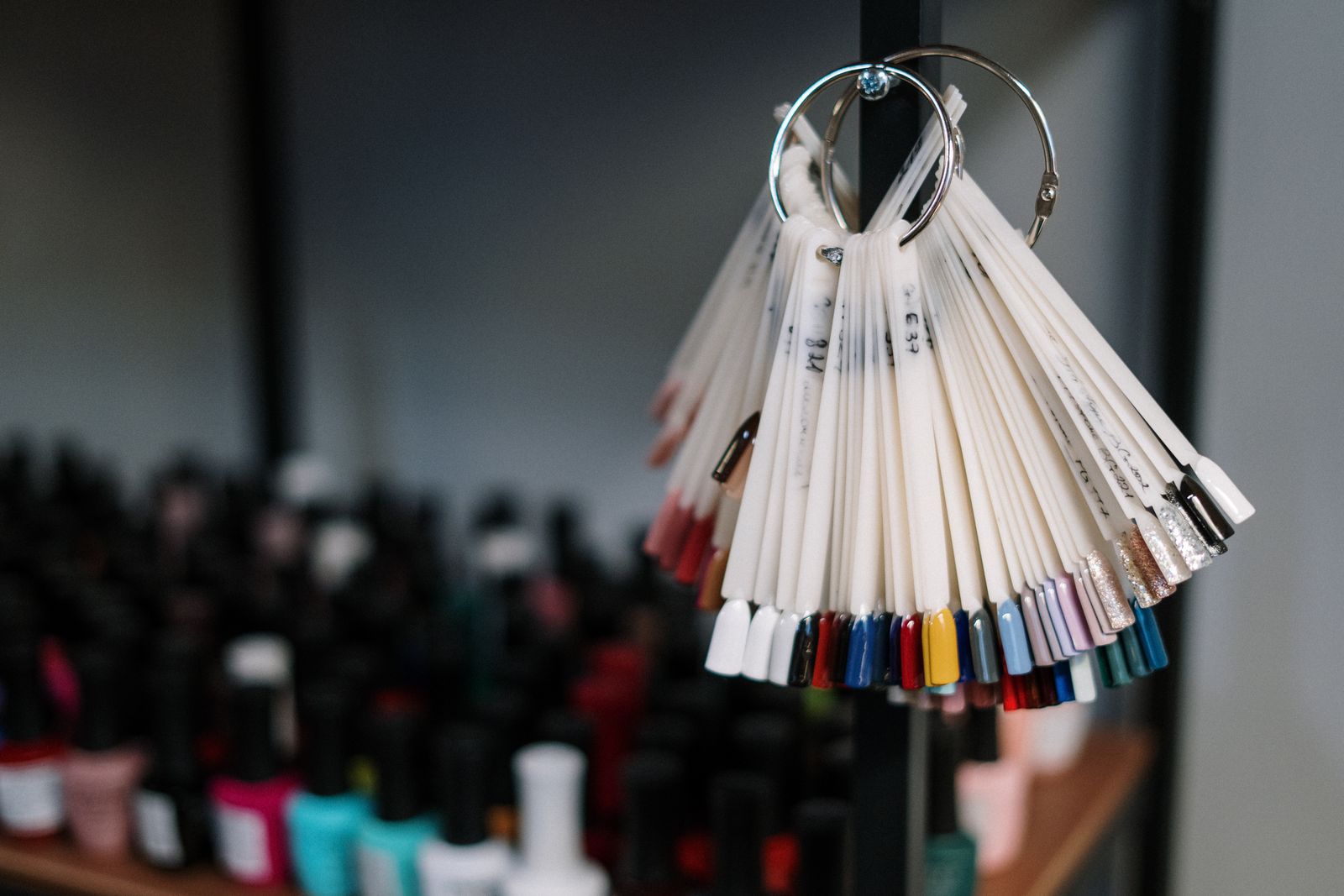 A pair of metal binder rings on a black shelf frame holding a fan of white nail-colour swatch sticks with a spectrum of coloured and glitter tips along the bottom edge; blurred rows of polish bottles on a wooden shelf behind.
