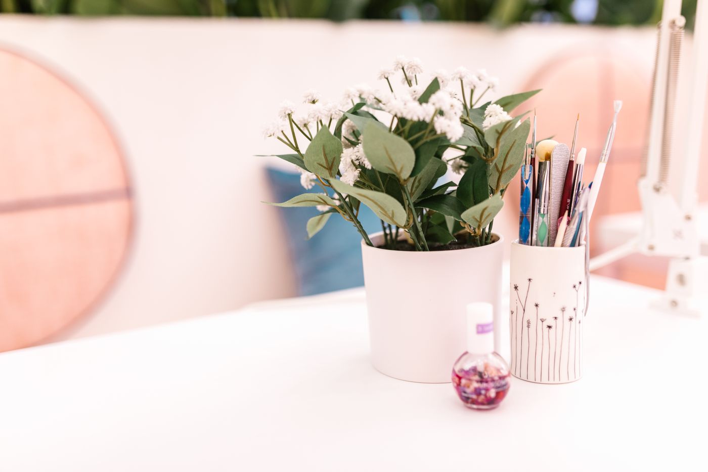 A white nail table with a white pot of small white flowers, a white ceramic cup holding nail-art brushes and files, a small round cuticle-oil bottle, and a white desk-lamp arm at right; blurred peach chair backs behind.