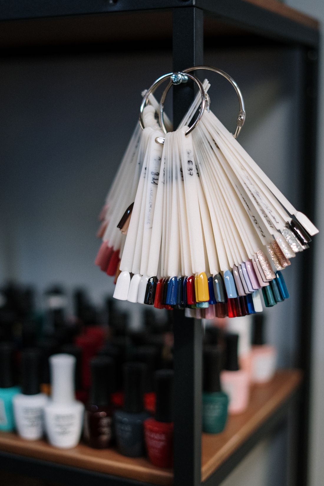 Nail-colour swatch rings shown vertically: tips in white, black, reds, blues, yellow, lilac and glitter gold and silver, with blurred gel-polish bottles on a wooden shelf below.